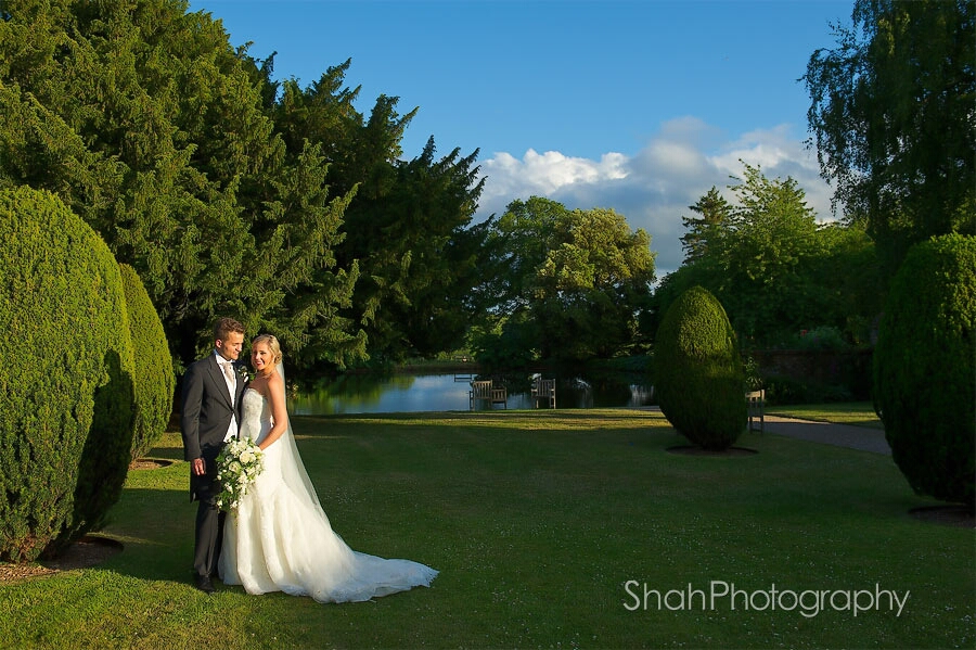 Bride and groom together standing in a pool of sunlight surrounded by trees with a background of blue sky and a lake