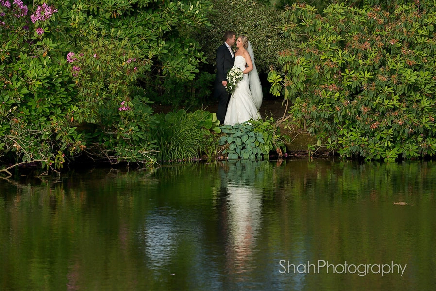 The bride and groom holding each other. A wide angle view with a foreground of a lake, their reflections in the water and a backdrop of evergreen bushes 
