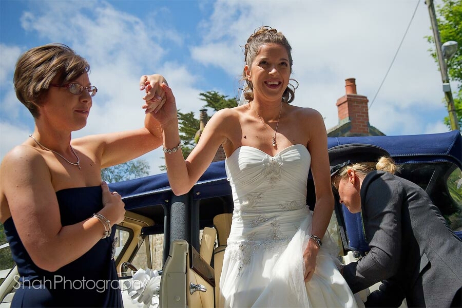 A low angle for the smiling bride getting out of her wedding car. The driver is helping with the dress and the bridesmaid is holding
the bride's hand